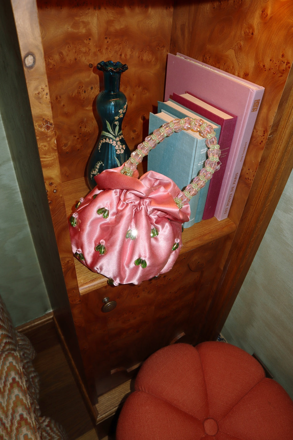 Pink satin Suryo bag with a beaded handle, styled beside books in a wooden cabinet